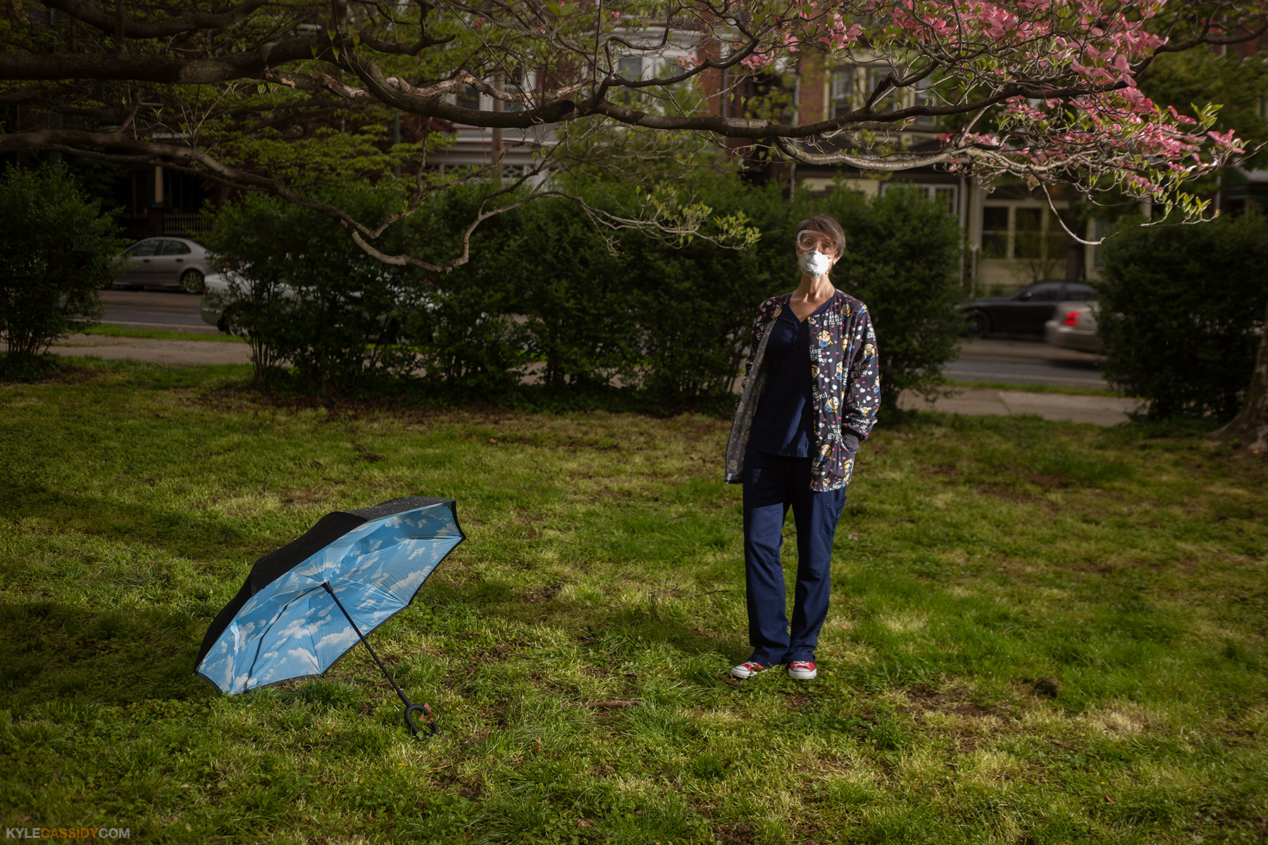 Karen wearing hospital scrubs standing in a park with an open umbrella on the ground next to her. She is wearing a surgical mask.