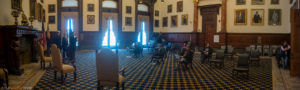 A large room in city hall with very few people in it, spread out in chairs 15 feet apart.